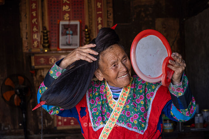 Elderly woman with long hair looking into a handheld mirror, showcasing unseen realities captured by a photographer.