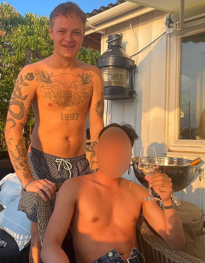 Norwegian Crown Princess’s son shirtless outdoors with tattoos, posing with another man holding a wine glass at sunset.