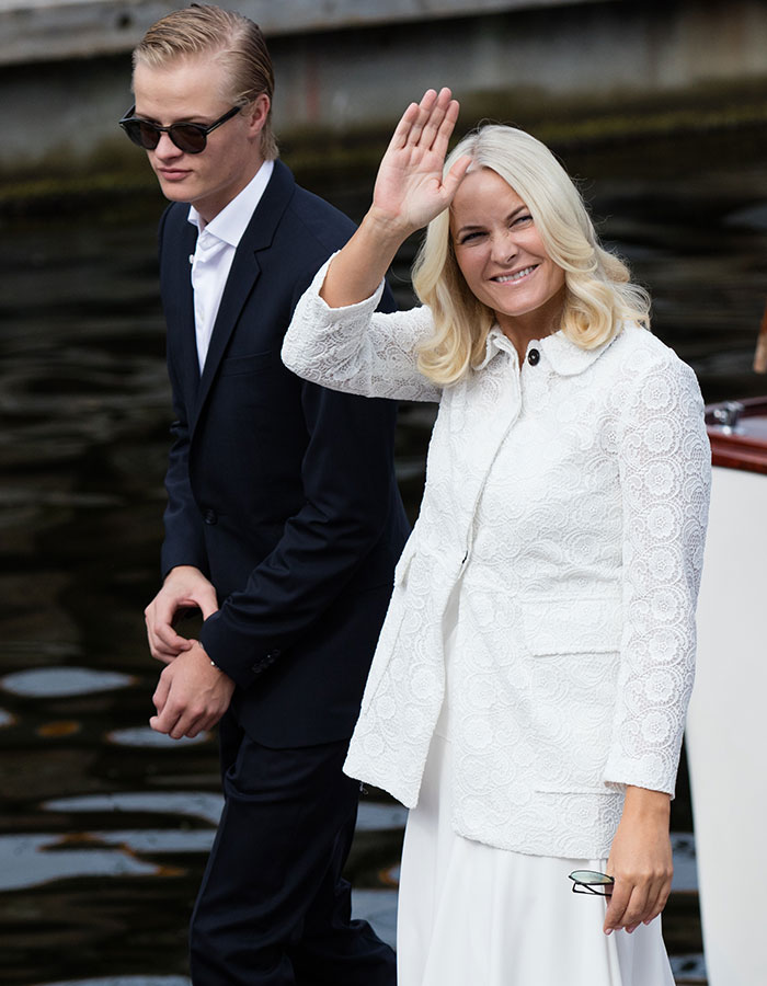Norwegian Crown Princess’s son in dark suit and sunglasses walking by the water while a woman in white waves and smiles.