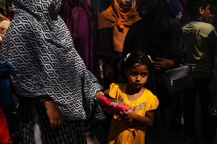 Child in yellow dress holding hands with woman in patterned hijab, candid street moment capturing emotion during travel.