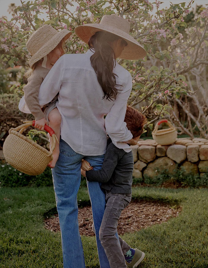 Woman in a hat carrying a child and holding hands with another child in a garden, related to narcissist Meghan Markle focus topic. Woman in a hat carrying a child and holding hands with another child in a garden, related to narcissist Meghan Markle focus topic.