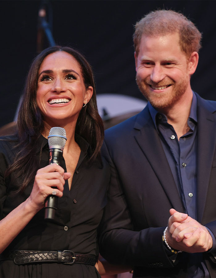 Meghan Markle holding a microphone and smiling beside Prince Harry during a public event with a black background. Meghan Markle holding a microphone and smiling beside Prince Harry during a public event with a black background.