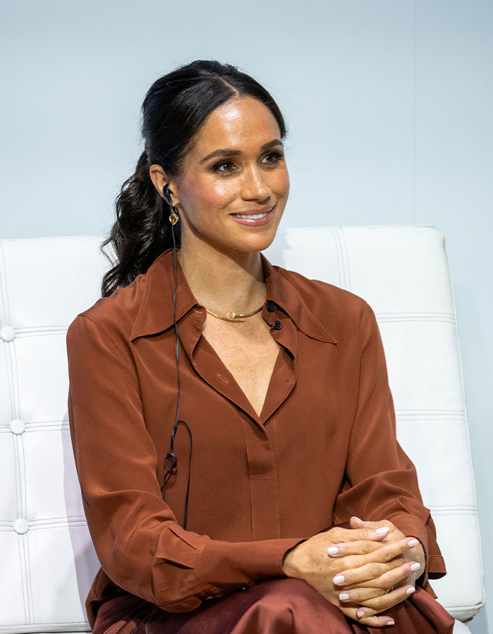 Meghan Markle seated in a brown outfit, wearing an earpiece and smiling during a public speaking event. Meghan Markle seated in a brown outfit, wearing an earpiece and smiling during a public speaking event.
