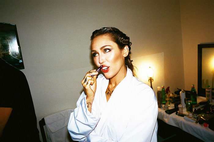 Miley Cyrus in a white robe, applying makeup backstage, highlighting her brutal infection and doctors speculating on the root cause. Miley Cyrus in a white robe, applying makeup backstage, highlighting her brutal infection and doctors speculating on the root cause.