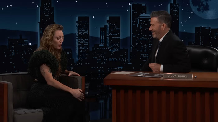 Miley Cyrus discussing her brutal infection with Jimmy Kimmel on a nighttime talk show set with city skyline backdrop. Miley Cyrus discussing her brutal infection with Jimmy Kimmel on a nighttime talk show set with city skyline backdrop.