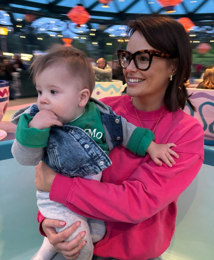 Woman wearing glasses holding a baby, smiling indoors, illustrating model with F-cup breast reduction surgery story.