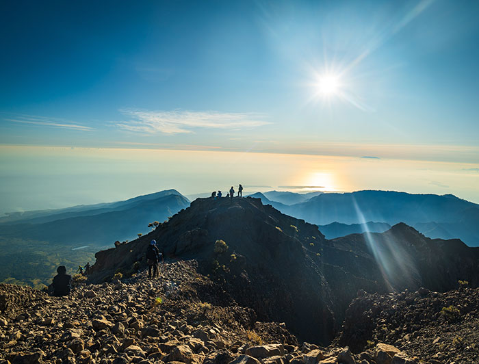 Hikers trekking near the summit of an Indonesian volcano under bright sun in a rocky mountainous landscape.