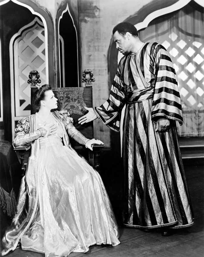 Actor in striped robe gestures while speaking to seated woman in period costume during Harlem Renaissance theater performance.