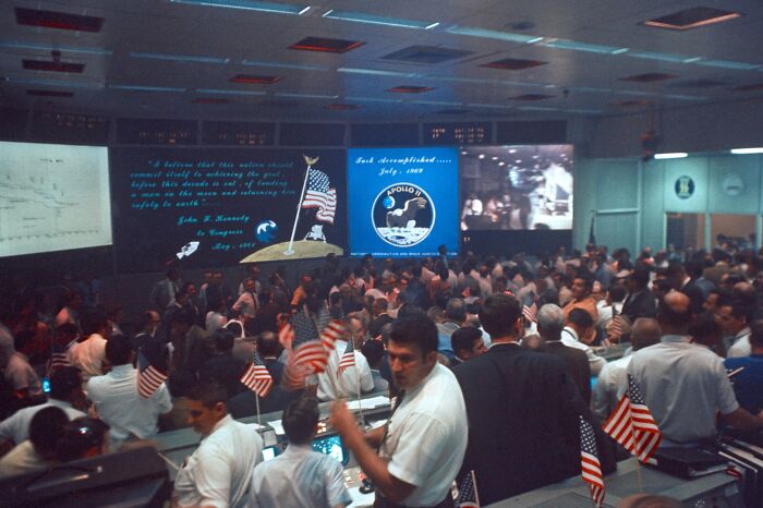 Mission control team celebrating Apollo mission success, waving American flags in a crowded room with historic screens and displays.