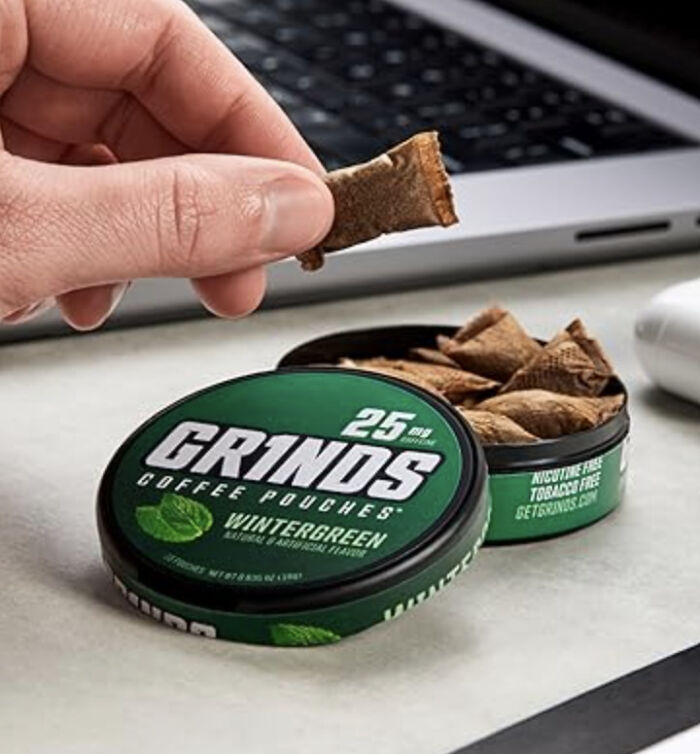 Hand holding a coffee pouch from a container of Grinds coffee pouches near a laptop, illustrating bizarre finds affecting finances.