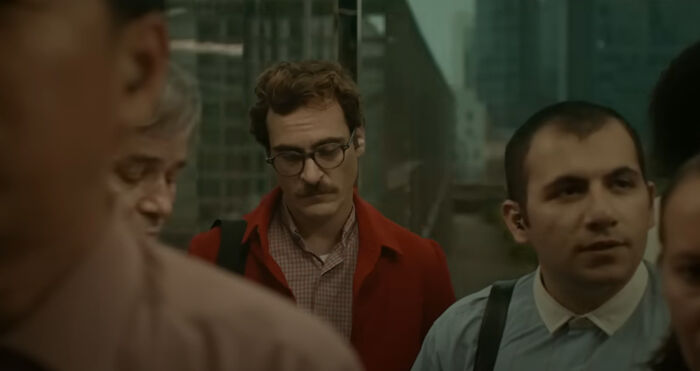 Man with glasses and mustache in red jacket on crowded elevator, a scene from one of the greatest films of the new millennium.