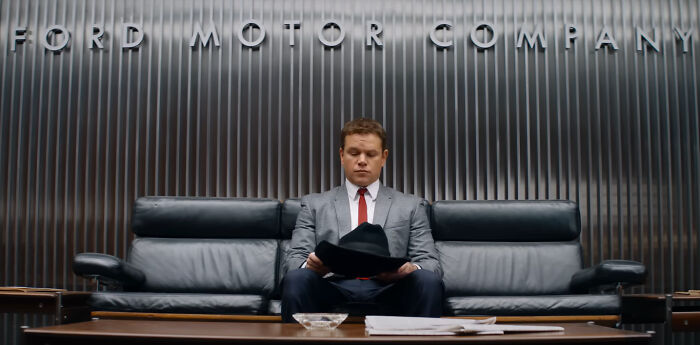 Man in a suit sitting on a black leather couch holding a hat, representing a scene from one of the greatest films of the new millennium.