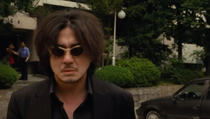 Man with sunglasses and messy hair in a dark suit standing outside in a scene from one of the new millennium's greatest films.