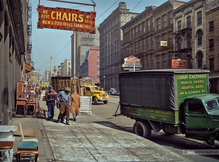 Colorized historical photo showing people and vintage trucks near a chair exchange sign on a city street.