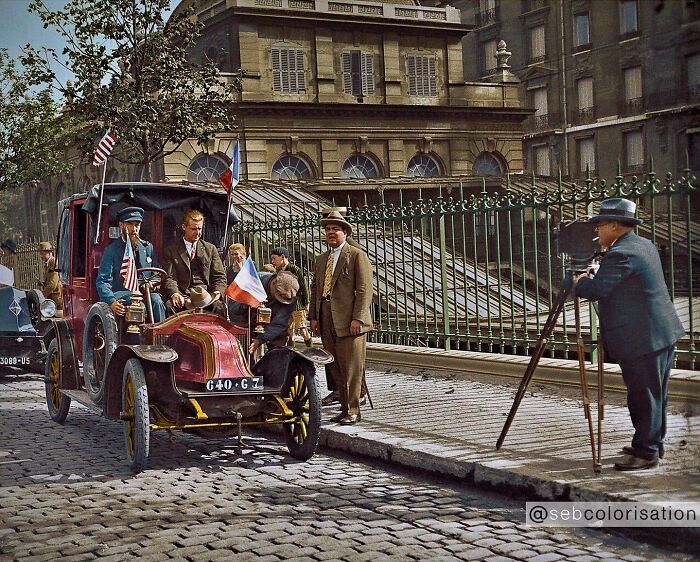 Early 1900s vintage car with American and French flags, photographed by a historical photos colorized artist on cobblestone street.