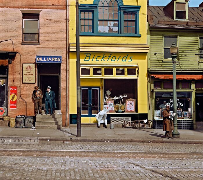 Colorized historical photo showing early 20th-century street scene with shops and people interacting outside.