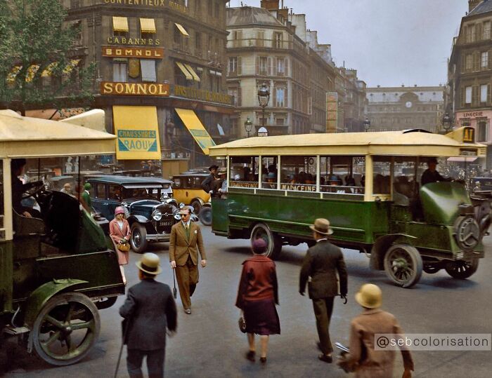 Colorized historical photo showing 1920s city street with vintage cars, tram, and people dressed in period clothing.