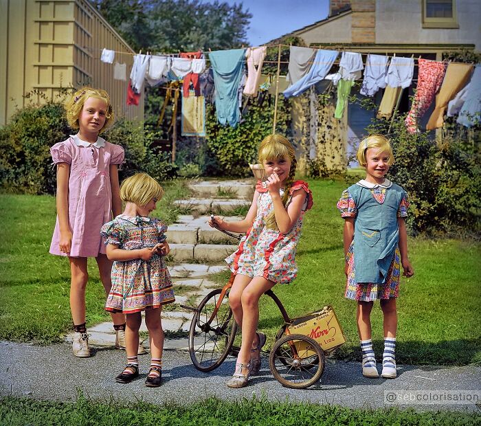 Four children in vintage dresses outside a house with colorful clothes hanging on a line, colorized historical photo.