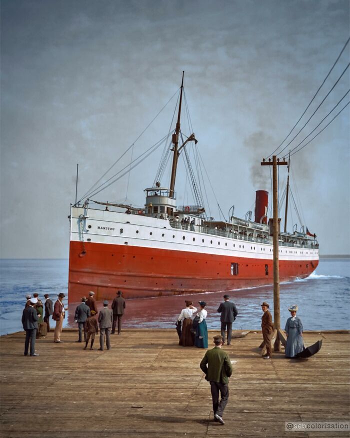 Colorized historical photo of a large red and white ship docked with people dressed in early 20th century clothing on the pier.
