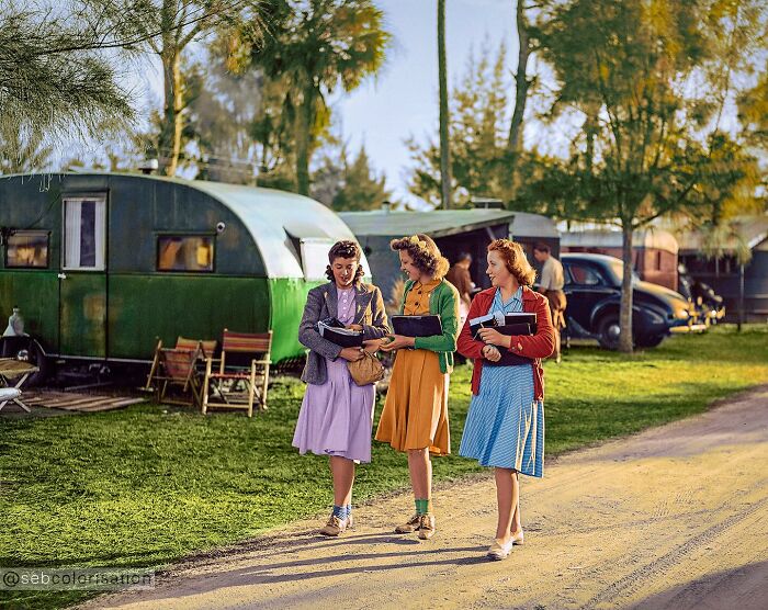 Three women in colorful vintage dresses walking with books near trailers in a magical historical photo colorized by a viral artist.