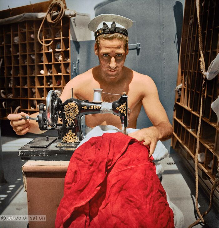 Colorized historical photo of a sailor using a vintage sewing machine to mend a large red fabric.