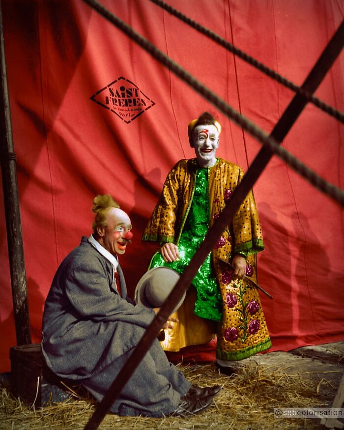 Two clowns in colorful costumes sitting and smiling against a red circus tent in colorized historical photo.