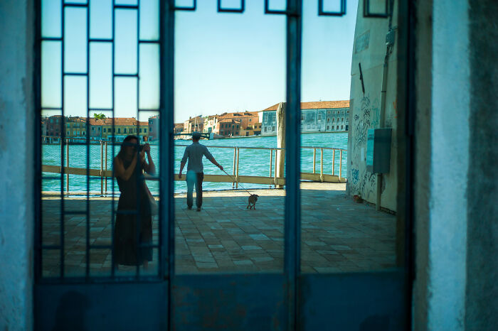 Reflection of a woman taking a photo and a person walking a dog by the water, cinematic photo capturing everyday life moments.
