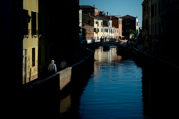 Cinematic photo of a canal with reflections and a person walking along the shadowed pathway beside old buildings.