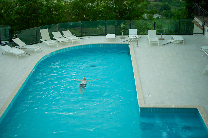 Person floating alone in a clear blue pool surrounded by empty lounge chairs in a cinematic everyday life photo.