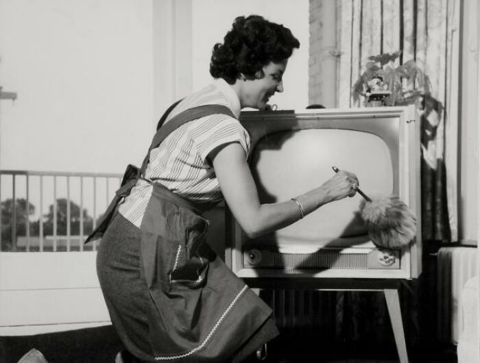 1940s housewife dusting an old television set wearing an apron in a vintage black and white home setting