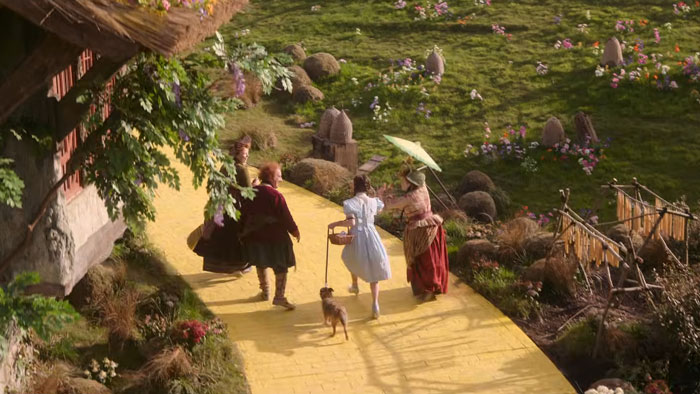 Scene from Wicked movie showing characters including Dorothy walking on the yellow brick road in a fantasy setting. Scene from Wicked movie showing characters including Dorothy walking on the yellow brick road in a fantasy setting.