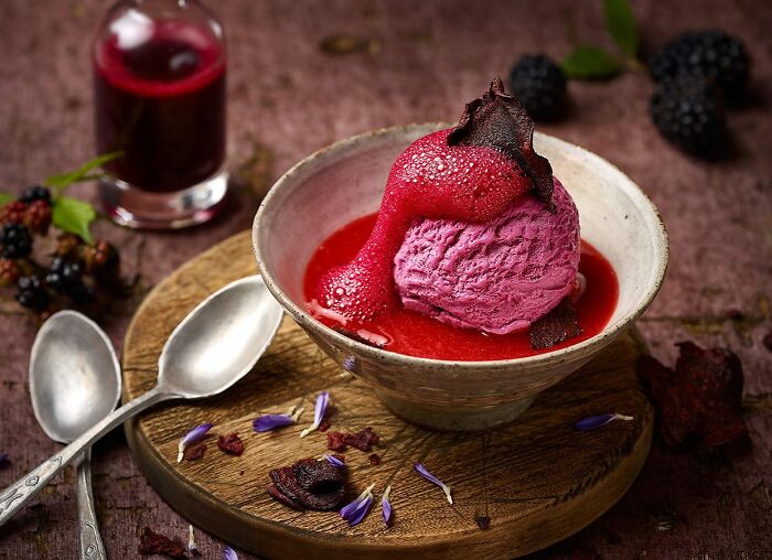 Vibrant mouthwatering food photo of a rich berry ice cream scoop with sauce in a bowl, styled with fresh blackberries and flowers.