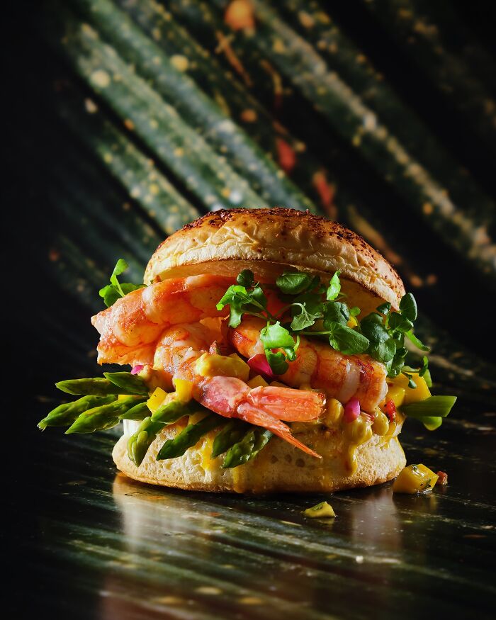 Shrimp sandwich with fresh greens, asparagus, and sauce on a toasted bun, showcasing mouthwatering food photography by Kai Stiepel.