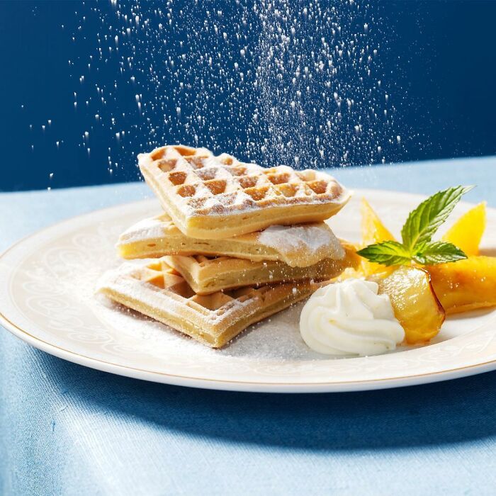 Stacked waffles dusted with powdered sugar, served with whipped cream and fruit, showcasing mouthwatering food photography.