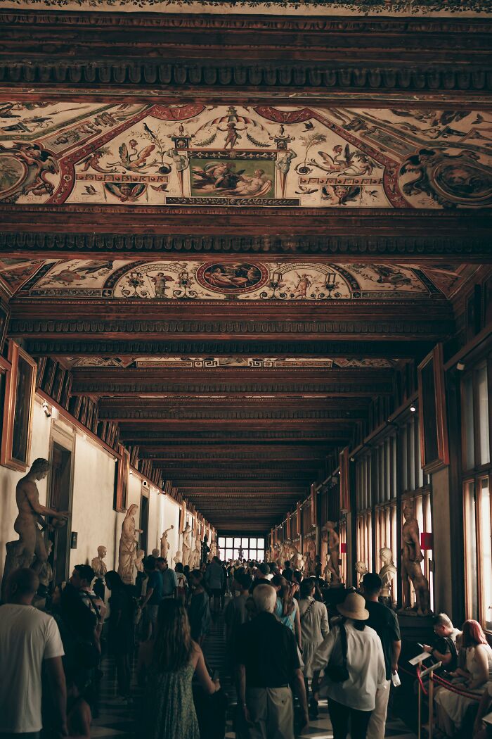 Crowded Italian museum hallway with tourists admiring 18th century painting and sculptures under ornate ceiling.