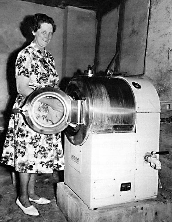 1940s housewife smiling while using a large vintage washing machine in a rustic indoor setting.