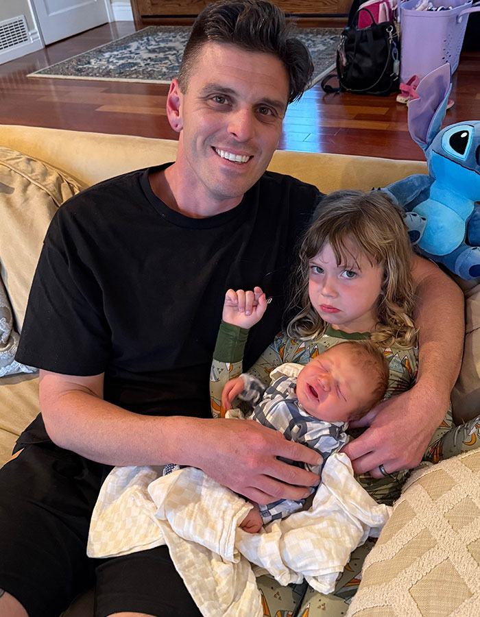 Man sitting on a couch holding two children, capturing a tender family moment after a real housewives exec childbirth loss. Man sitting on a couch holding two children, capturing a tender family moment after a real housewives exec childbirth loss.