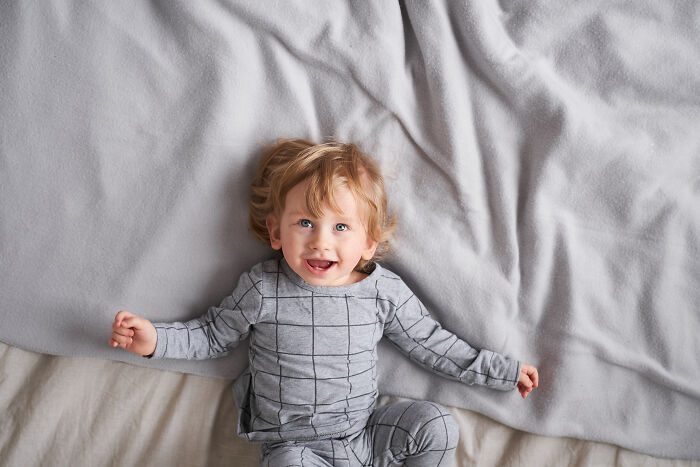 Smiling toddler lying on a bed wearing gray checkered pajamas, representing baby names that secretly weird people out.