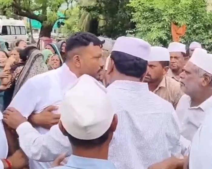 Sole survivor of Air India crash embraced by men in white caps during emotional gathering outdoors near trees and vehicles. Sole survivor of Air India crash embraced by men in white caps during emotional gathering outdoors near trees and vehicles.