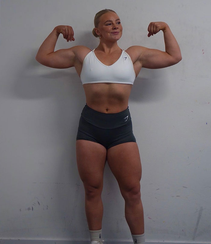 Woman powerlifter flexing muscles in white sports bra and black shorts against plain wall, showcasing strength and fitness. Woman powerlifter flexing muscles in white sports bra and black shorts against plain wall, showcasing strength and fitness.