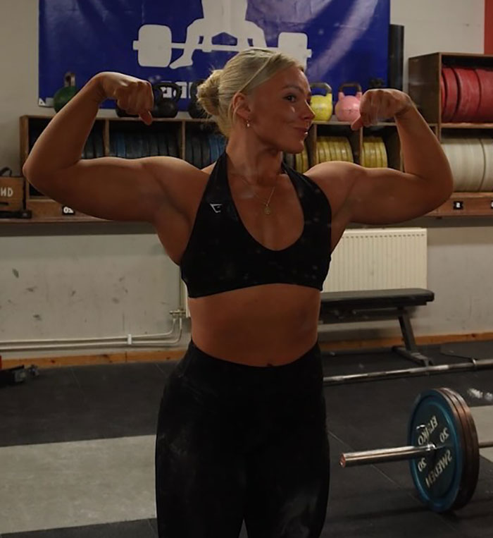 Female powerlifter flexing muscles in gym with barbell, related to viral video of woman urinating during competition. Female powerlifter flexing muscles in gym with barbell, related to viral video of woman urinating during competition.