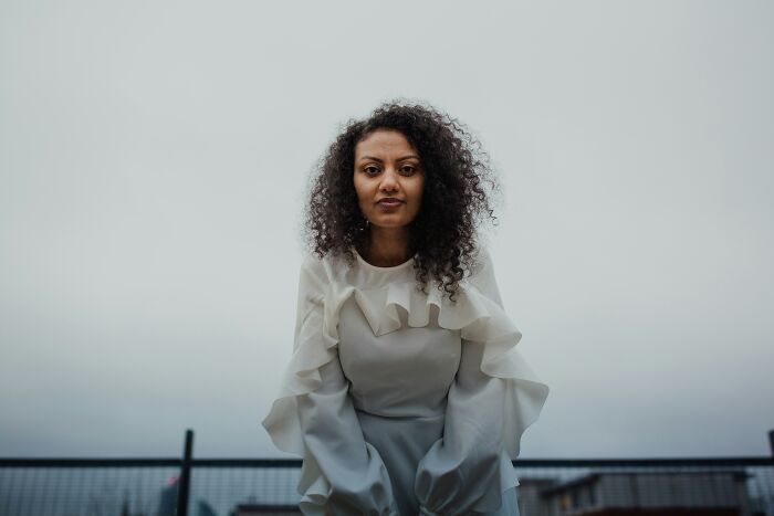 Woman with curly hair and white blouse showing subtle signs of low self-esteem in an outdoor setting with overcast sky.