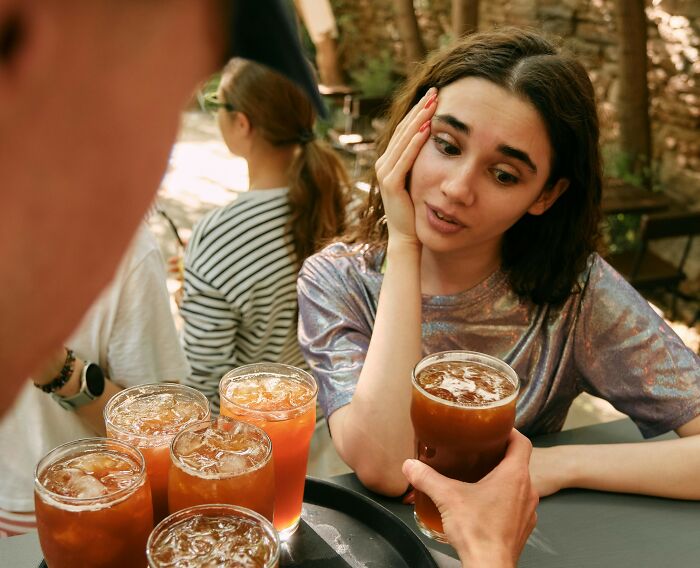 A woman on a date looking frustrated as a bartender serves a tray of drinks at an outdoor bar setting.