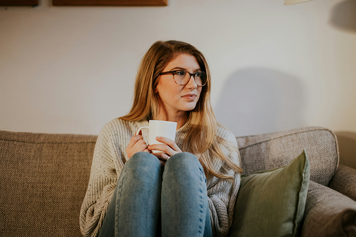 Young mom sitting on couch holding a white mug, looking thoughtful in a cozy home setting.