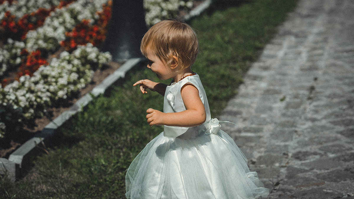 Toddler girl in a white dress walking outside at a family wedding, highlighting young mom and illegitimate kid situation.