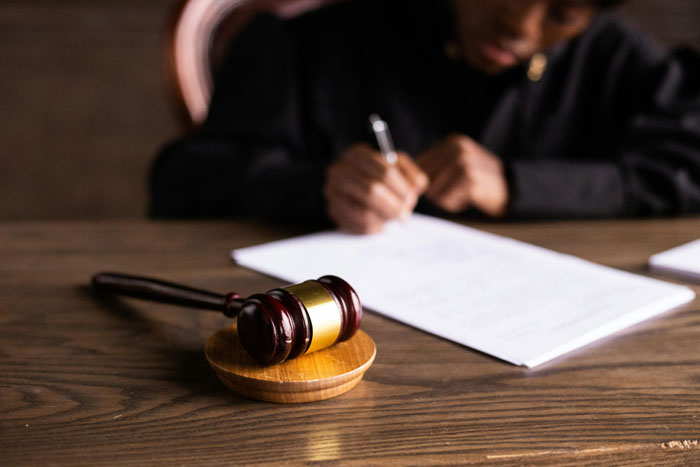 Close-up of a wooden gavel with a person writing legal documents in the background about MIL, pregnant DIL, and divorce. Close-up of a wooden gavel with a person writing legal documents in the background about MIL, pregnant DIL, and divorce.