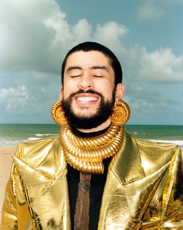 Bad Bunny smiling on the beach wearing a gold metallic jacket and large gold jewelry reflecting his net worth and fashion style.