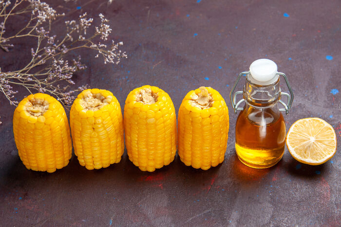 Four pieces of corn arranged beside a bottle of oil and a lemon half with dried flowers on a dark surface representing bad things people life.