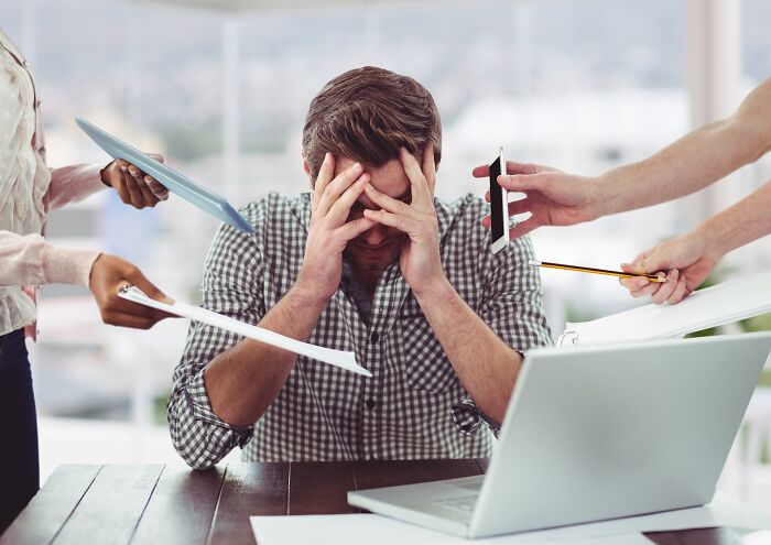 Stressed man overwhelmed with work, surrounded by hands offering papers, phone, and pencil.