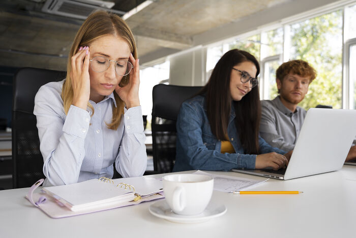 Woman stressed at work, holding her head while colleagues work on a laptop nearby.